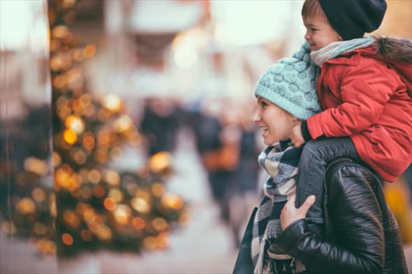 Une mère porte son fils sur ses épaules et se baladent devant un sapin de Noël illuminé.