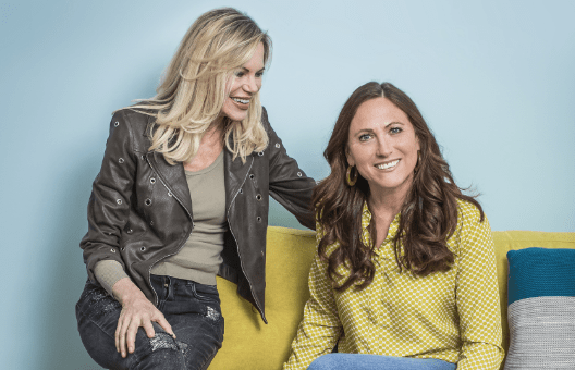 Nina Ruge und Sennerin Martina Fischer sitzen auf einem gelben Sofa und lächeln. Auf der blauen Wand hinter ihnen sieht man das Victoria-Logo.